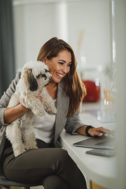 Foto de una joven mujer de negocios sentada con su perro mascota y usando en su computadora portátil durante el trabajo desde casa. - Foto, Imagen