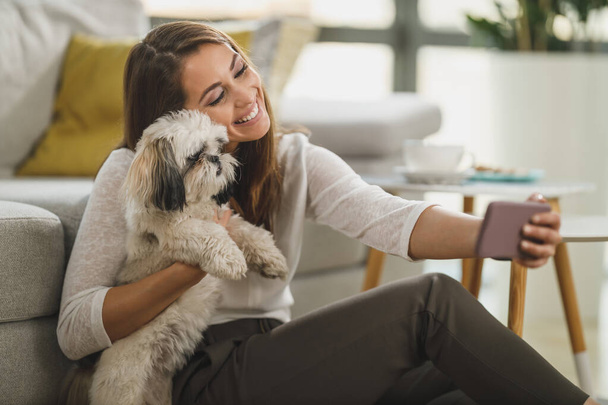 Laukaus söpö nuori nainen ottaen ja selfie rentoutuen lemmikkikoiran kanssa olohuoneessaan kotona. - Valokuva, kuva