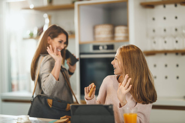 Съемка милой девочки-подростка и ее улыбающейся матери приветствуют друг друга у себя дома, в то время как мама готовится к работе. - Фото, изображение