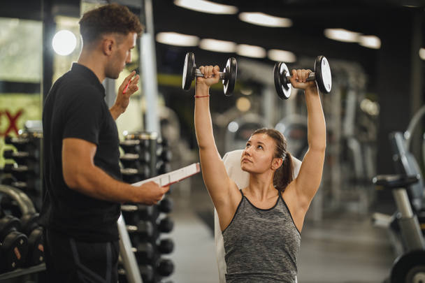 Shot of a muscular young woman in sportswear working out with personal trainer at the gym machine. She is pumping up her muscule with dumbbell. - Photo, Image