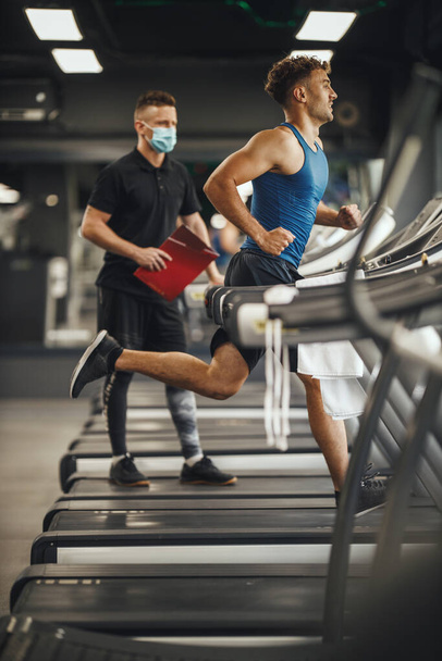 Una foto de un joven musculoso corriendo en la cinta en el gimnasio. Está trabajando con un entrenador personal.. - Foto, Imagen
