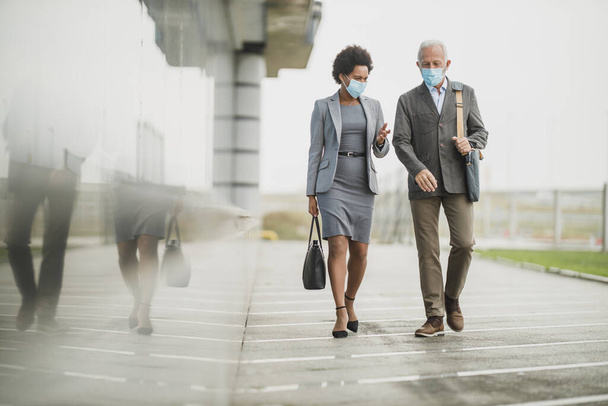 COVID-19パンデミック時のオフィスビル前を歩きながら、保護マスクを持つ2人の成功した多民族ビジネスの人々のショット. - 写真・画像