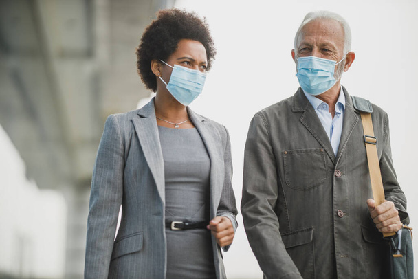 Shot of two successful multi-ethnic business people with protective mask having a discussion while walking in front of the office building during COVID-19 pandemic. - Photo, Image