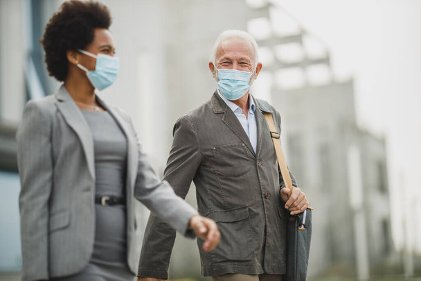 Fotografía del exitoso empresario senior y su colega negra usando máscara protectora y hablando mientras caminaba frente al edificio de oficinas durante la pandemia de COVID-19. - Foto, Imagen