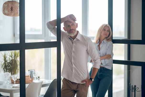 Bearded man looking sad, blonde woman watching him with arms crossed - Foto, imagen