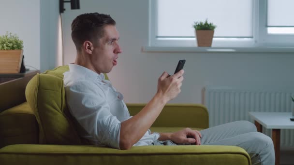 Excited overjoyed man winner celebrating success looking at smartphone and making yes gesture while sitting at home on couch. Mobile victory concept - Footage, Video