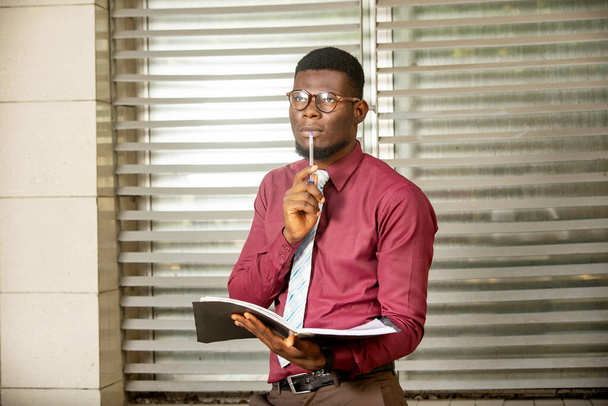 joven empresario africano en cachemira de manga larga y gafas sosteniendo documento y pluma y mirando a la pared. - Foto, imagen