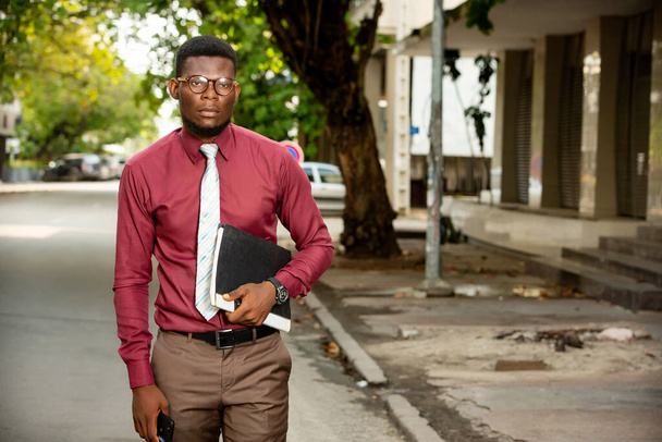 Όμορφος Αφρικανός που περπατάει στο δρόμο κρατώντας έγγραφα. φορώντας ένα πολύ κομψό μπορντό κόκκινο πουκάμισο φορώντας γραβάτα και γυαλιά - Φωτογραφία, εικόνα