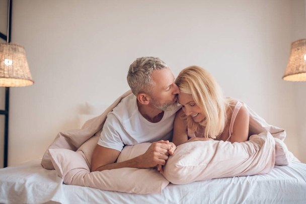 Grey-haired man and a blonde woman lying on the bed and looking romantic - Fotó, kép