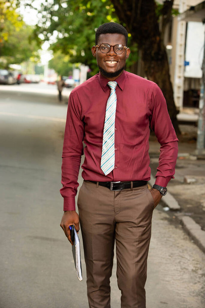 Foto de un joven y hermoso africano caminando al aire libre sosteniendo un cuaderno en su brazo vestido con pantalones marrones y una elegante chemise roja borgoña usando gafas y una corbata - Foto, imagen