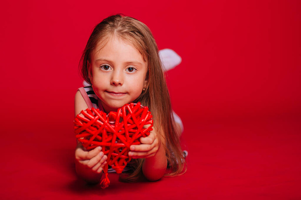 Een klein meisje met blond haar in een gestreepte jurk houdt een rood hart in haar handen. - Foto, afbeelding