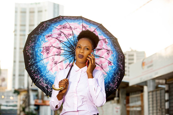 hermosa mujer joven sosteniendo un paraguas y hablando en el teléfono celular mientras camina por la calle - Foto, imagen