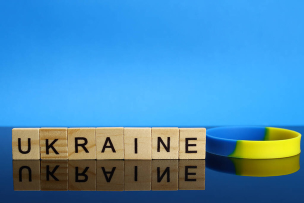 Ukraine Wort aus Holzbuchstaben und einem gelb-blauen Armband auf einer Spiegeloberfläche - Foto, Bild