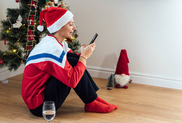 Mujer joven del milenio usando el teléfono en casa en Nochebuena. Compras de vacaciones en línea, cuarentena celebración virtual  - Foto, Imagen