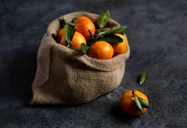 Mandarins with green leaves. Bright orange tangerines with leaves lie in a burlap sack on a dark background. Christmas fruit. Dark photo. The photo shows a textured background - Fotoğraf, Görsel
