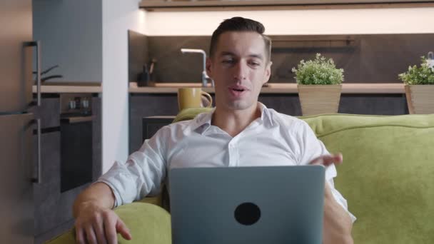 Egy fiatalember videochat-et használ a laptopján, hogy kommunikáljon a barátaival vagy üzleti partnereivel. Jóképű férfi beszél, és nézi laptop képernyőn, miközben ül a kanapén otthon nappali - Felvétel, videó