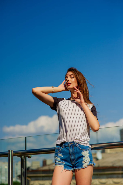 Dívka v džínových kraťasech na balkóně. Letní portrét mladé stylové hipster dívka pózující na balkóně - Fotografie, Obrázek