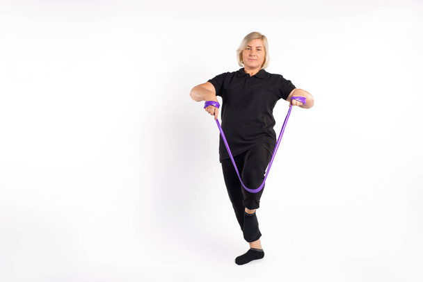 Senior woman stretches fitness elastic band with her leg. White background with empty space. Healthy lifestyle and pandemic concept. High quality photo - Fotografie, Obrázek