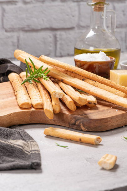 Традиционные итальянские хлебные палочки гриссини с розмарином, сыром пармезан, оливковым маслом, чесноком и солью на сером фоне. - Фото, изображение