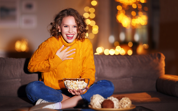 mladá potěšená veselá žena jíst popcorn směje se a sleduje vánoční komediální film na kabelovou televizi doma ve večerních hodinách alon - Fotografie, Obrázek