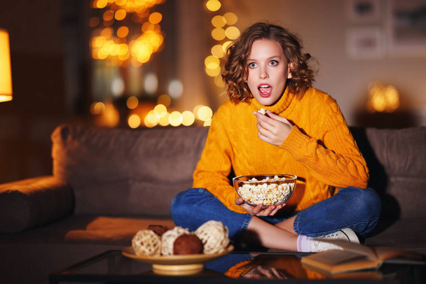 Γοητευμένη νεαρή γυναίκα σε πουλόβερ τρώει ποπ κορν και βλέποντας ενδιαφέρουσα ταινία στην τηλεόραση, ενώ κάθεται στον καναπέ το βράδυ στο hom - Φωτογραφία, εικόνα