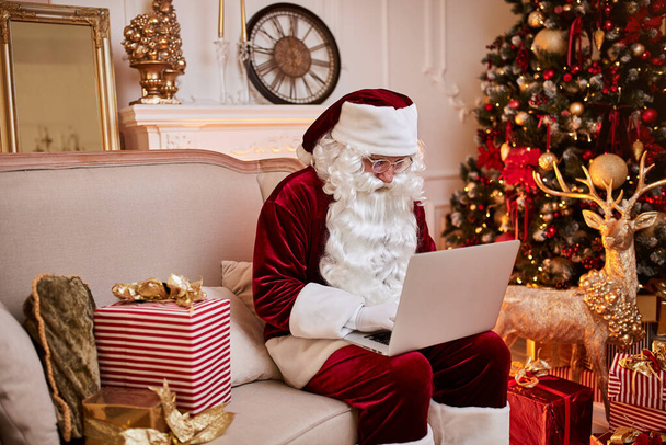 Joulupukki istuu kotonaan ja lukee sähköpostia kannettavan tietokoneen kanssa joulu pyytää tai toivelista lähellä takka ja puu lahjoja. Uusi vuosi ja Hyvää joulua, hyvää joulua käsite - Valokuva, kuva