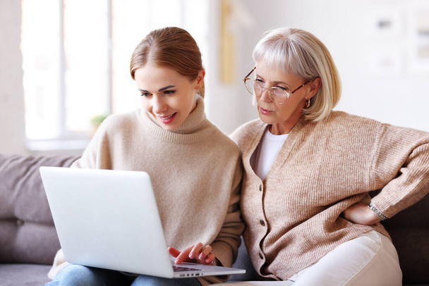 Erwachsene Frau lehrt konzentrierte ältere Mutter, Laptop zu benutzen, während sie auf der Couch im gemütlichen Wohnzimmer zusammen sitzt - Foto, Bild