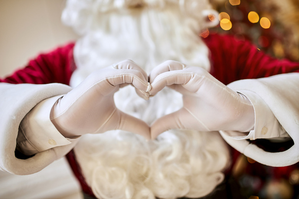 Santa Claus forming shape of heart with her fingers near the fireplace and christmas tree with gifts. New year and Merry Christmas , happy holidays concept - Φωτογραφία, εικόνα