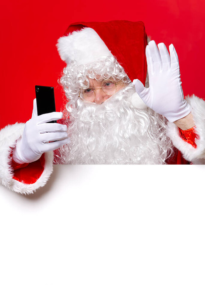 Santa Claus holding mobile phone in hand while standing behind the blank white board. copy space. Santa Claus texting. - Valokuva, kuva
