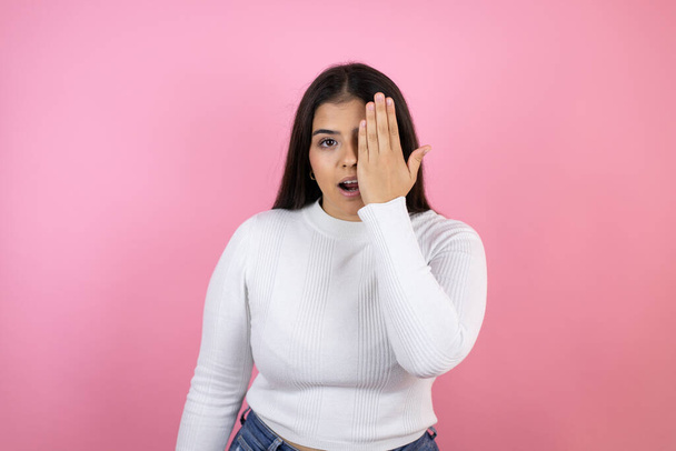 Young beautiful woman over isolated pink background covering one eye with hand, confident smile on face and surprise emotion. - Photo, Image