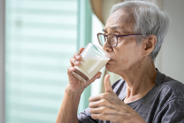 Lähikuva, aasialainen vanhempi nainen juo tuoretta maitoa lasista, terveet vanhukset, joilla on lasi maitoa, vanhukset, jotka näyttävät peukaloa juomisen aikana, terveydenhuolto, terveellinen ravitsemus, ruoka ja juomat käsite - Valokuva, kuva