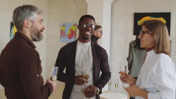 PAN tiro de jovens convidados multiétnicos masculinos e femininos segurando copos de champanhe, sorrindo e conversando na festa de abertura da exposição na galeria de arte - Filmagem, Vídeo