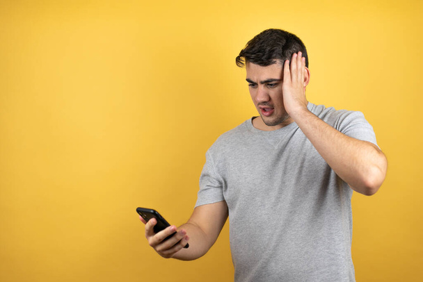 Fiatal jóképű férfi visel alkalmi póló felett elszigetelt sárga háttér kezében egy telefont meglepett arckifejezéssel - Fotó, kép