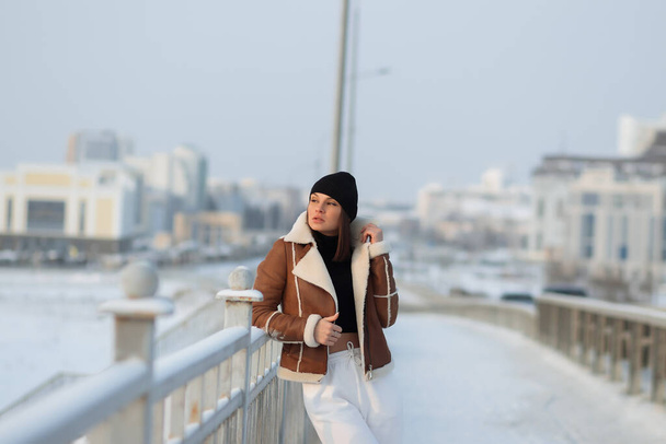 Intrygująca kobieta w brązowej kurtce z owczej skóry z kapeluszem i patrząc na kamerę stojącą na śnieżnym moście miejskim - Zdjęcie, obraz