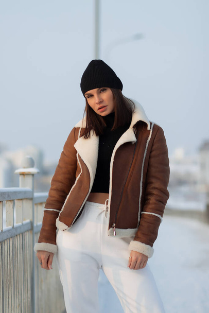 Mulher sedutora vestindo casaco de pele de carneiro marrom com chapéu e olhando para a câmera em pé na ponte da cidade nevada - Foto, Imagem