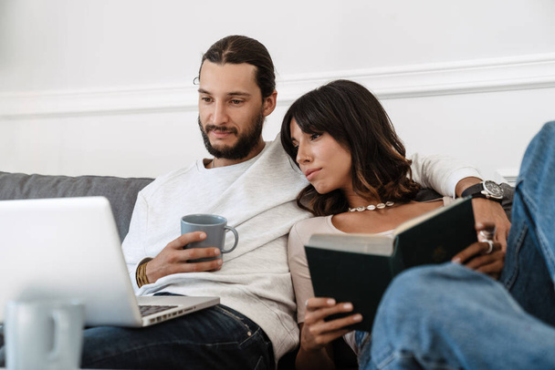 Image of focused beautiful couple reading book and using laptop while sitting on sofa at home - Foto, Bild