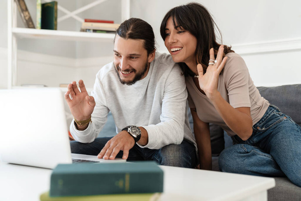 Image of cute cheerful couple using laptop and waving hands while sitting on sofa at home - Photo, Image