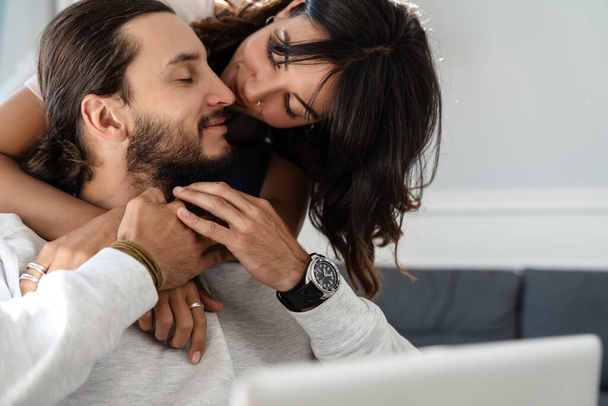 Image of cute happy couple using laptop and hugging while sitting on sofa at home - Photo, Image
