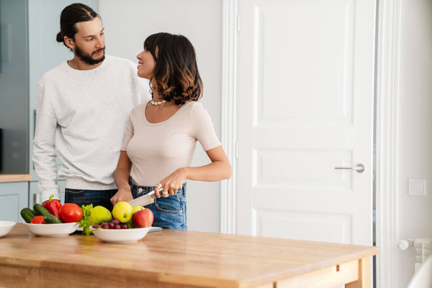 Image of cute happy couple preparing lunch while looking at each other in kitchen - Фото, изображение