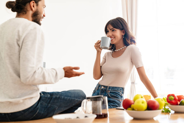 Image of joyful caucasian couple talking while drinking coffee at home kitchen - Foto, imagen