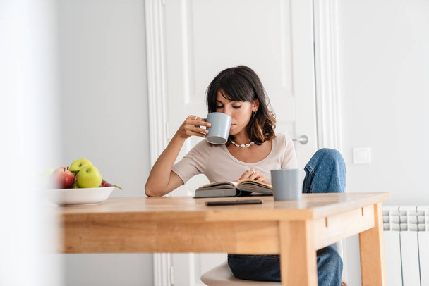 Image of serious beautiful woman reading book while drinking coffee at home kitchen - Foto, Imagem