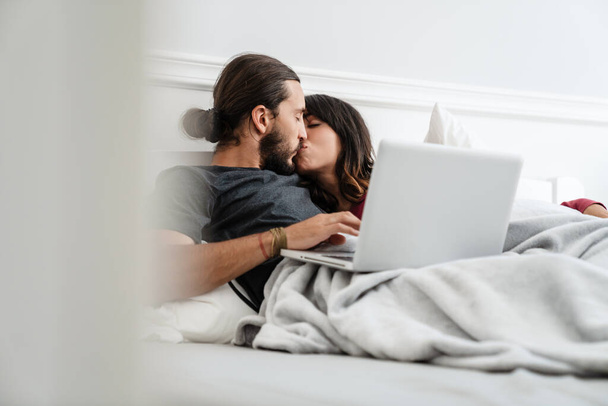 Image of romantic happy couple using laptop and kissing while lying in bed at home - Fotó, kép