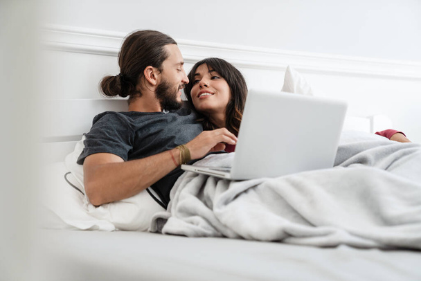 Image of romantic cheerful couple using laptop and hugging while lying in bed at home - Foto, Imagem