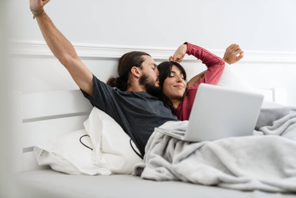 Image of romantic cheerful couple stretching their bodies while lying with laptop in bed at home - Photo, Image