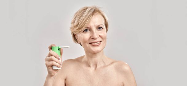 Portrait of attractive middle aged woman smiling at camera and holding throat spray, standing isolated over grey background - Foto, Imagem