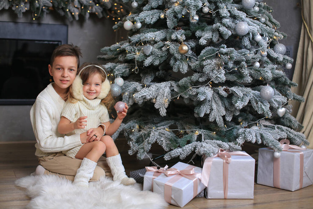 Los niños - el hermano en el jersey blanco de punto y la hermana en el vestido blanco de punto y los auriculares blancos de piel adornan el árbol de Año Nuevo. - Foto, imagen