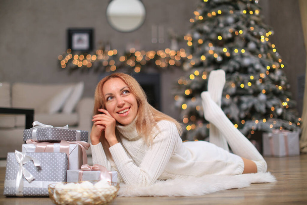 Ein weißes schönes Mädchen in weißem Strickkleid und Socken liegt auf einer Kunsthaut mit weißem Fell in der Neujahrsdekoration. Schachteln mit Geschenken liegen in der Nähe, es gibt einen Korb mit Marshmallows - Foto, Bild
