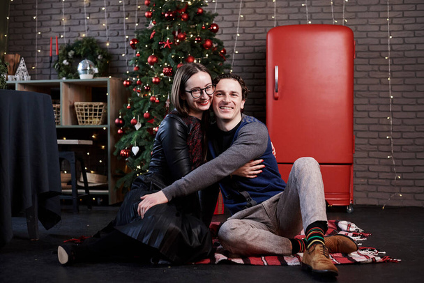 Young married couple sitting on kitchen floor in front of red fridge and Christmas tree. Man and woman celebrating winter holidays at home during quarantine at home. Happy new year and Merry Christmas - Foto, imagen