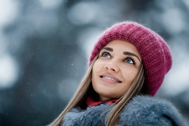 灰色の毛皮のコートと雪の日に冬の森の中を歩く赤い帽子のかわいい女の子の肖像画。松林の中の冬の気分。クリスマスの時間屋外. - 写真・画像