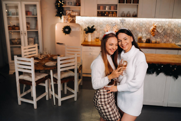 two girls in a cozy home environment with champagne in their hands at Christmas. Smiling girls drink champagne on a festive evening. - Foto, Imagem